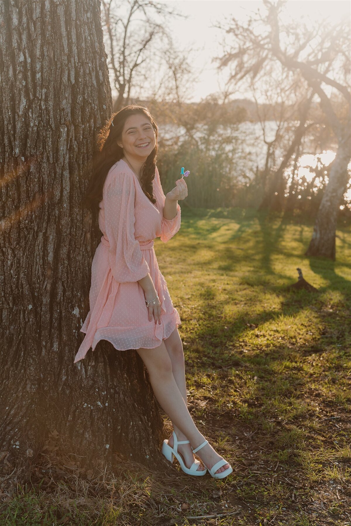 Teen girl in a light pink dress leaning against a tree with sunlight streaming through the leaves at Walter Hall Park in League City, TX. Captured during a Sweet 16 portrait session by ShutterUp Photography.