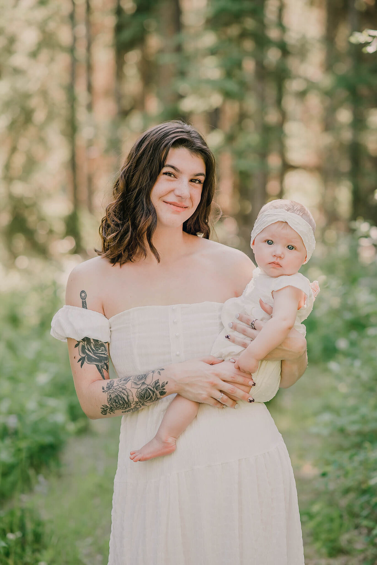 Mother holds baby daughter on her hip while in the forest. They both wear white and look at the camera. Taken by Brittany Danielle Photography.