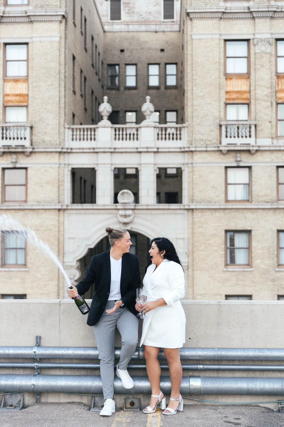Same-sex couple celebrating their engagement on a rooftop in downtown Dallas, popping champagne with city skyline in the background