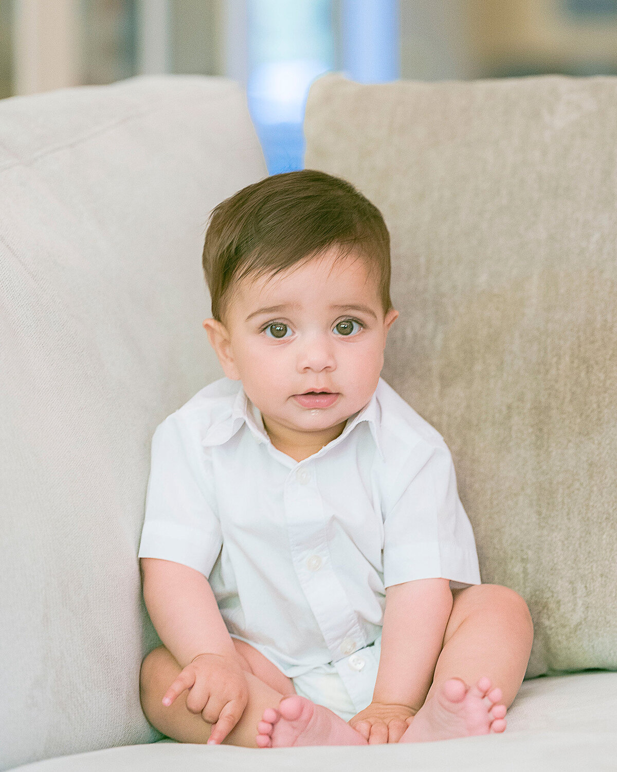 Seven month old baby wearing a white onsie sitting on a couch lit by natural light looking at the camera captured at home by San Francisco Newborn Photographer.