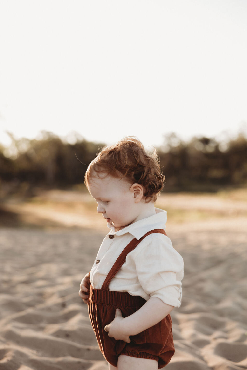 A young child with curly hair stands on sandy terrain, wearing a white shirt and reddish-brown suspenders, looking down thoughtfully—a moment beautifully captured by an NSW family photographer.