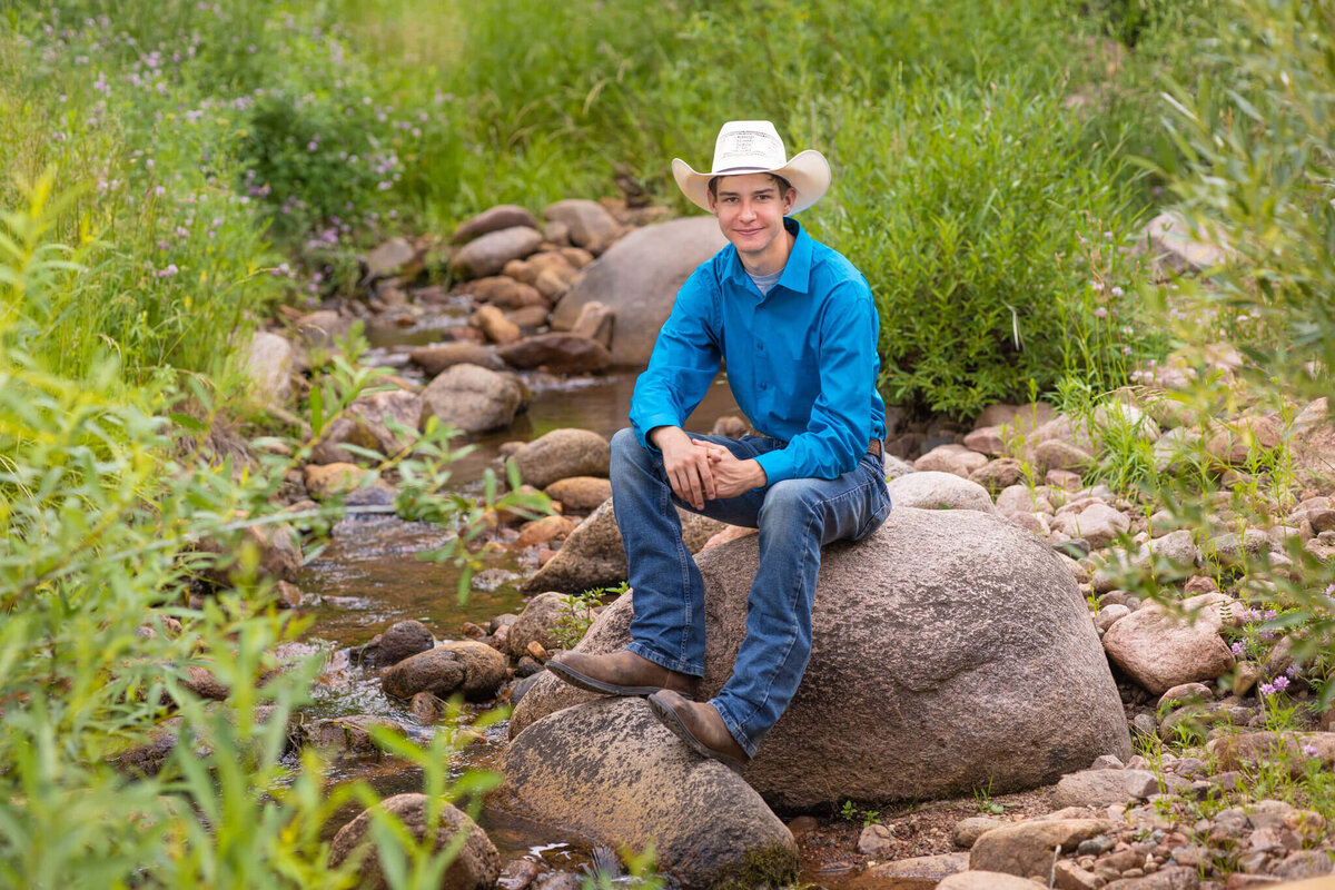 high school senior boy in a blue shirt and jeans wearing a cowboy hat sitting on a rock in a shallow creek