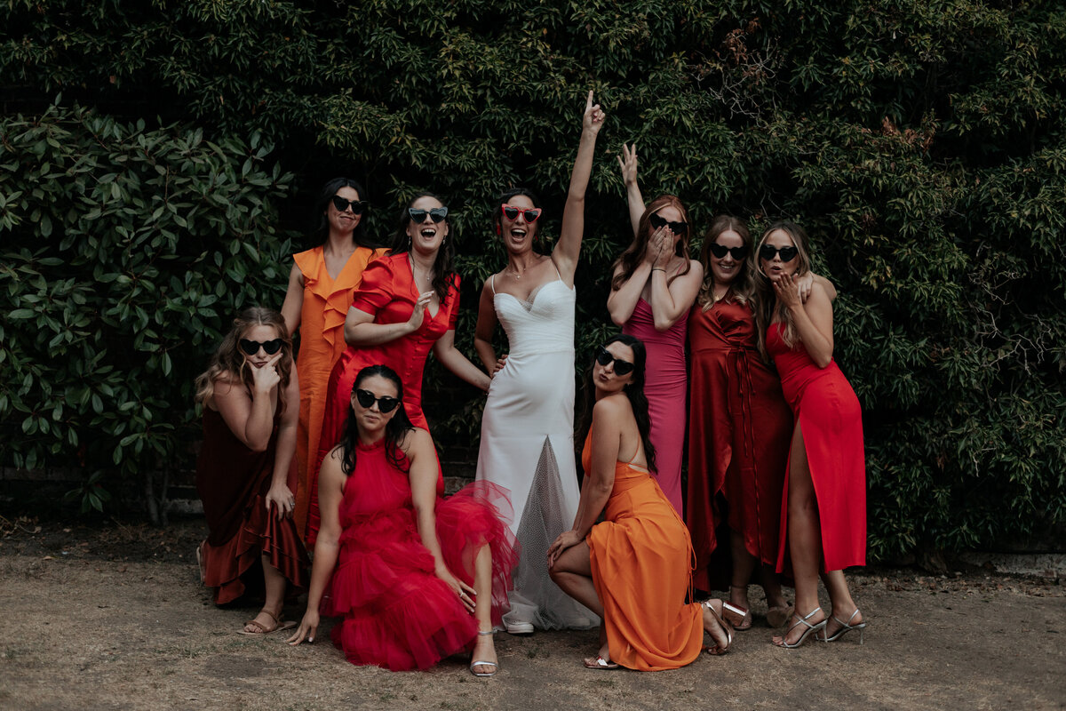 A bridal squad of mismatched red, pink and orange dresses pose around a bride wearing matching sunglasses