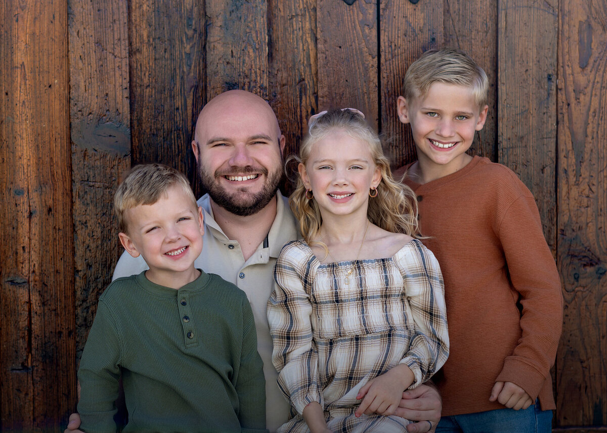 Dad and his 3 kids sit together in front of a wooden wall for their Iowa Family Portraits