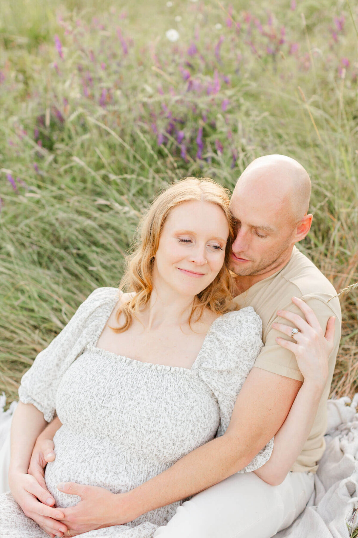 Pregnant woman sitting in front of husband and leaning back into him. They are holding her belly and he is nuzzled into her temple during their Portland sunset maternity photo session.