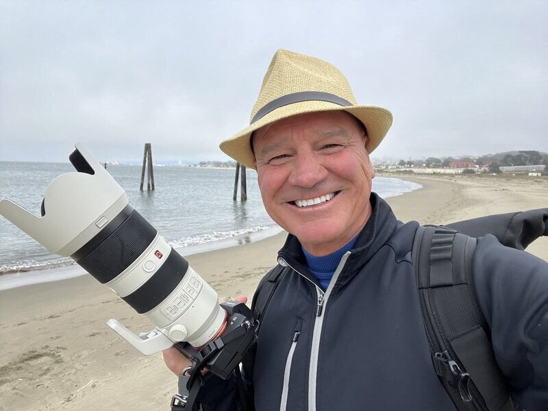 Man smiling on the beach wearing a fidora and a dark top while holding a camera with a very long lens