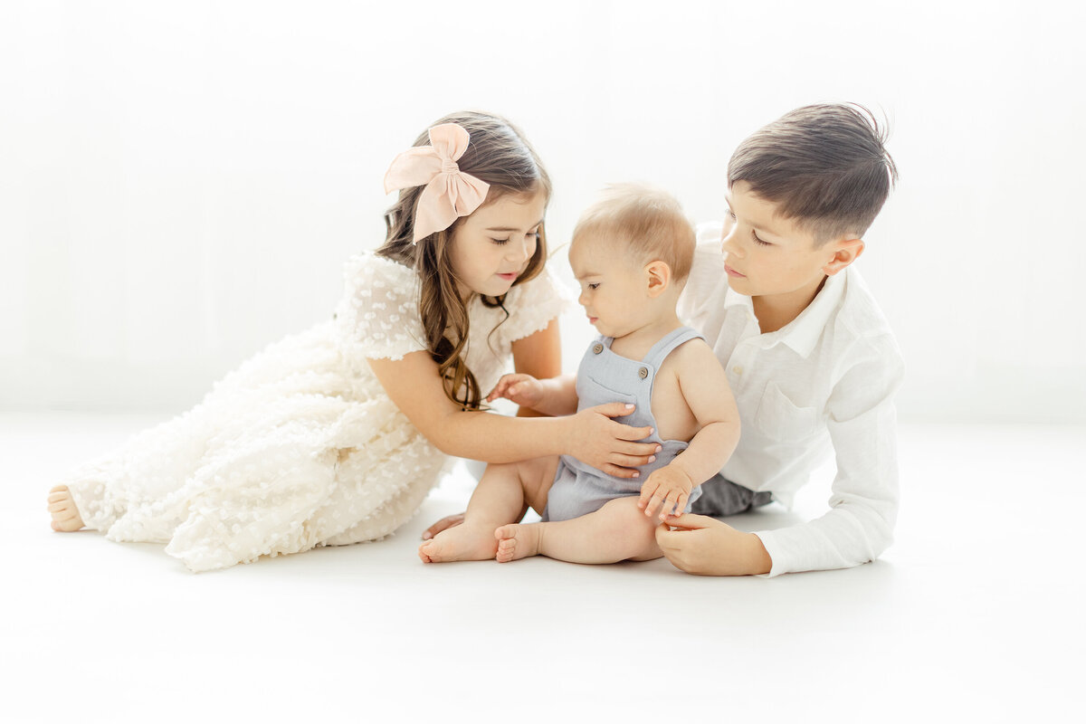 Siblings playing with their baby brother on a photography studio floor in Dallas for his milestone photos.