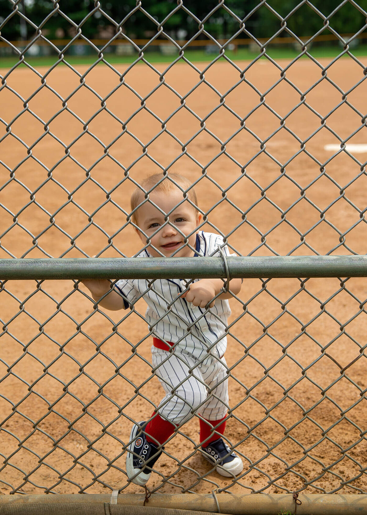 1 year old boy looking through the fence from out on the baseball diamond.