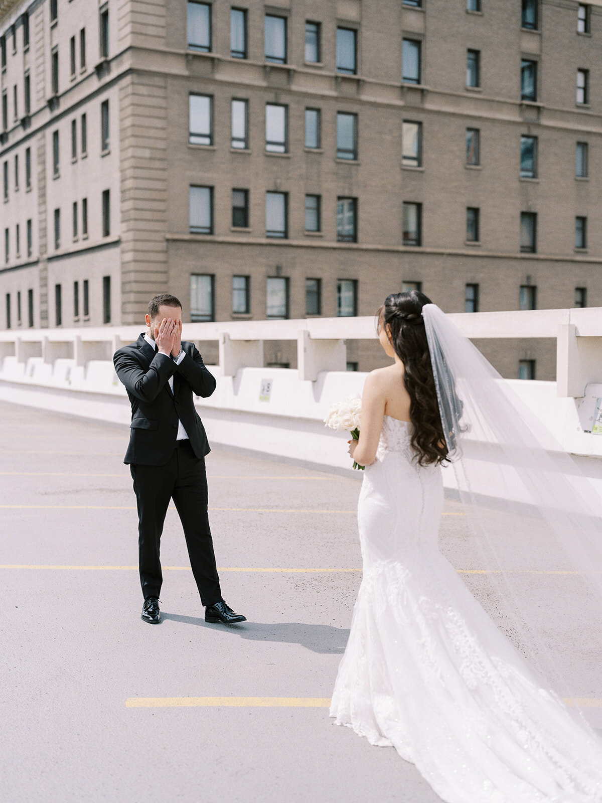 A groom in a black suit and bow tie covers his face with his hands while a bride in a white wedding dress and veil stands in front of him on a rooftop parking lot, capturing a beautiful moment from their classic Calgary wedding.