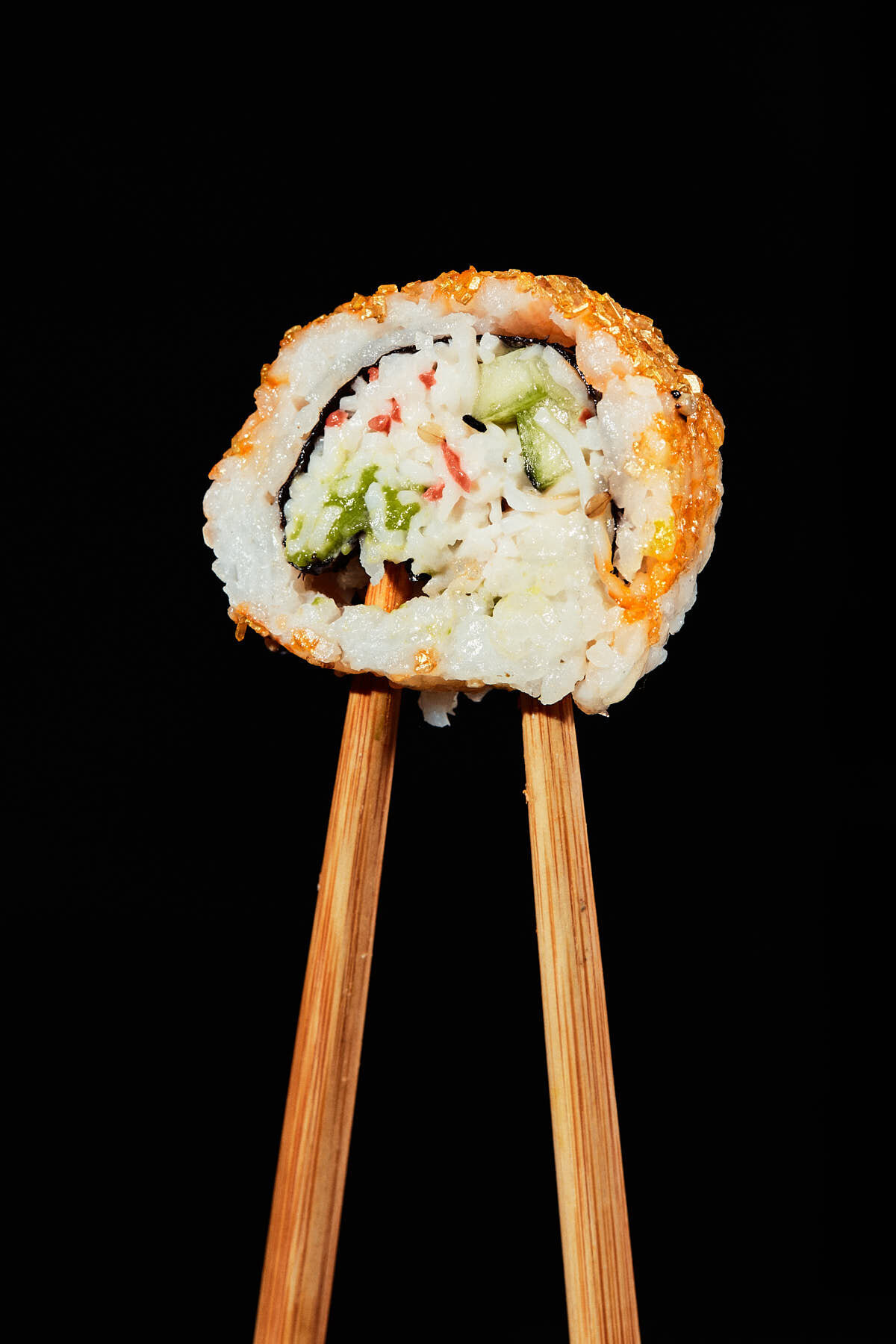 A close-up image of a sushi roll with cucumber, rice, and fish roe, held by chopsticks against a black background. Captured by Austin-based food photographer Mica McCook, this shot highlights the detailed textures and vibrant ingredients in sushi.