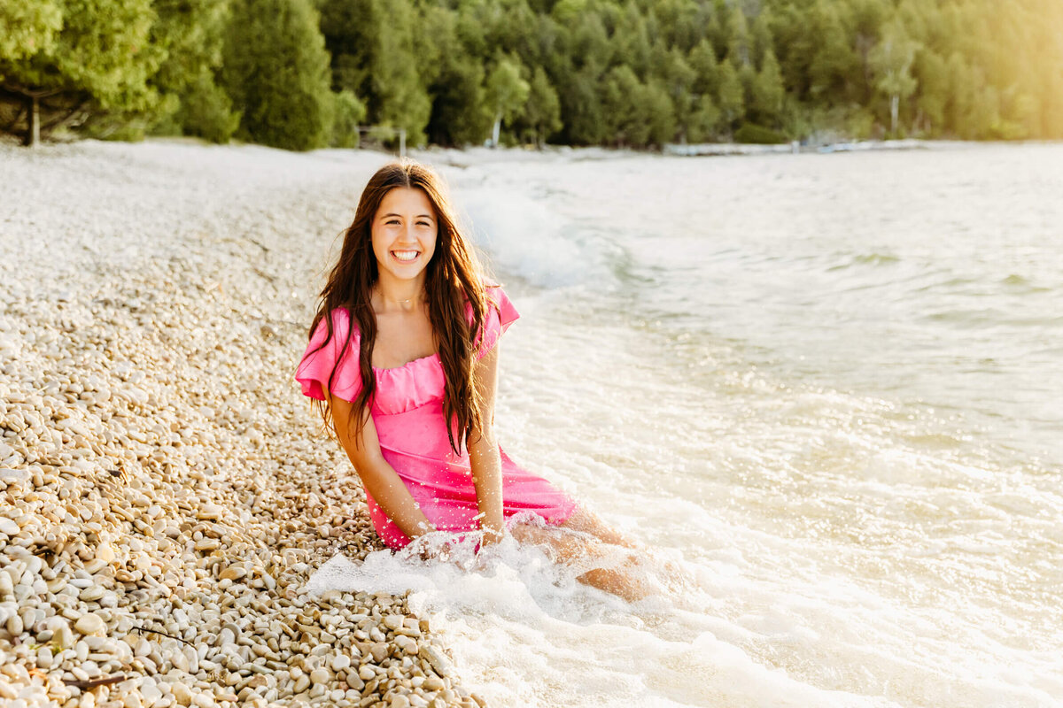 gorgeous teenage girl in a bright pink dress sitting on a white rock beach in Door County as a wave crashes over her