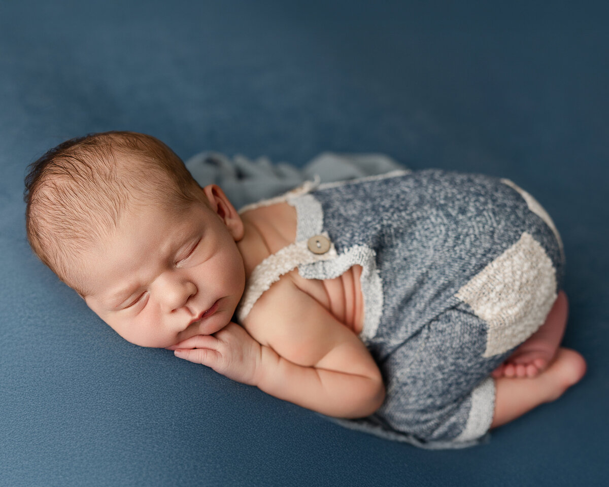 Newborn baby boy sleeping peacefully on a soft blue blanket, dressed in a textured blue romper with ivory knit trim. Captured by Inland Empire newborn photographer Kaitlyn Dawn Photography.