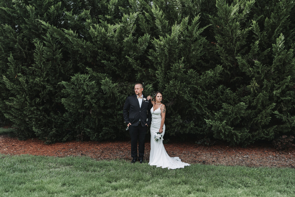 Bride and groom pose confidently in front of towering evergreens at The Gentry Farm wedding venue in Danville, Virginia.