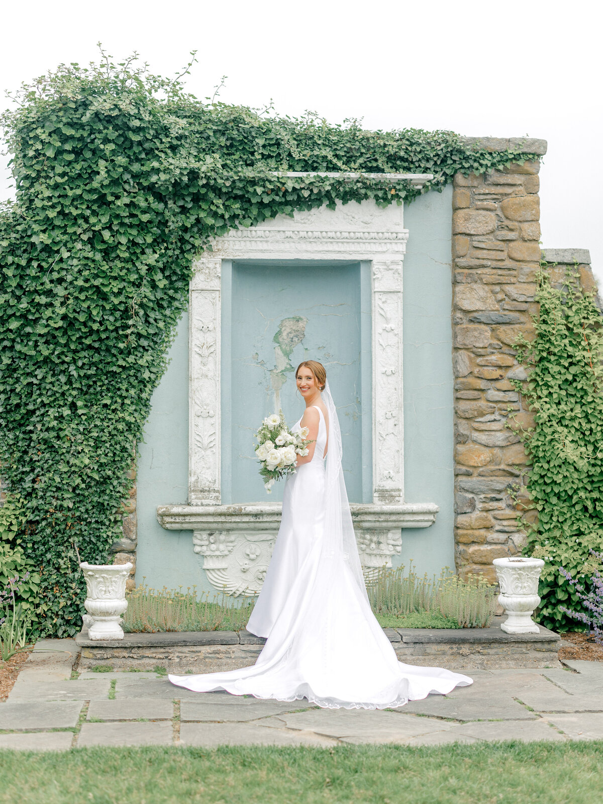Bride standing with her dress train behind her in front of a blue marble and stone wall covered in ivy