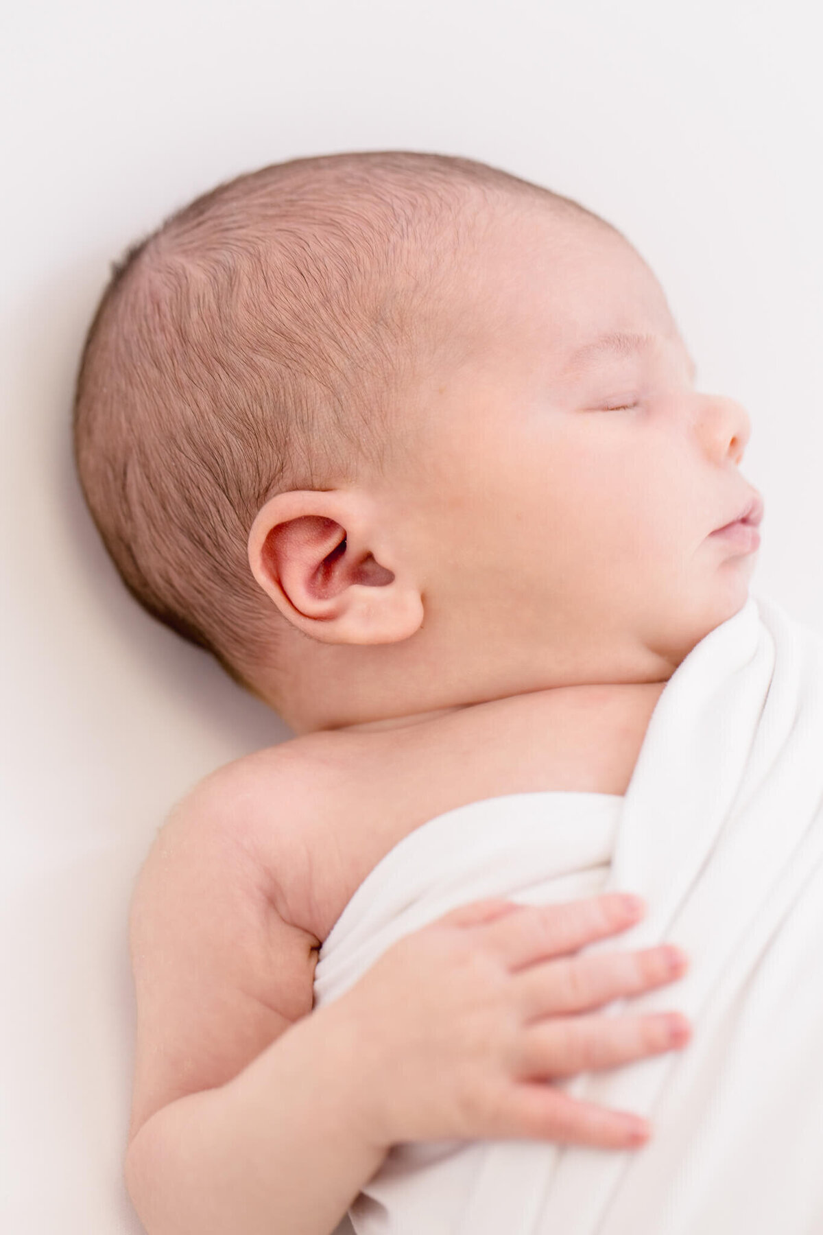 A newborn baby peacefully sleeping, wrapped in a soft white blanket. The babys head is turned to the side, with eyes closed and tiny fingers visible on the blanket. The background is light and softly blurred.