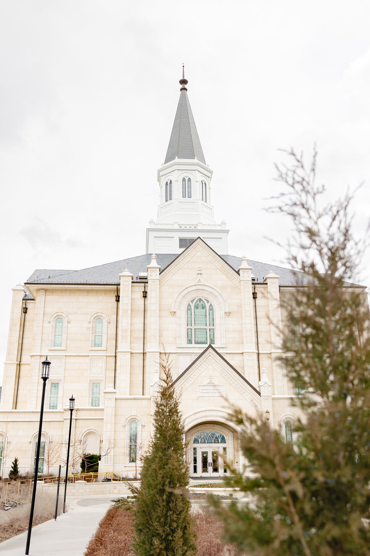 The front exterior of the Taylorsville Utah LDS Temple with a tall spire and light stone facade, framed by winter trees on a cloudy day.