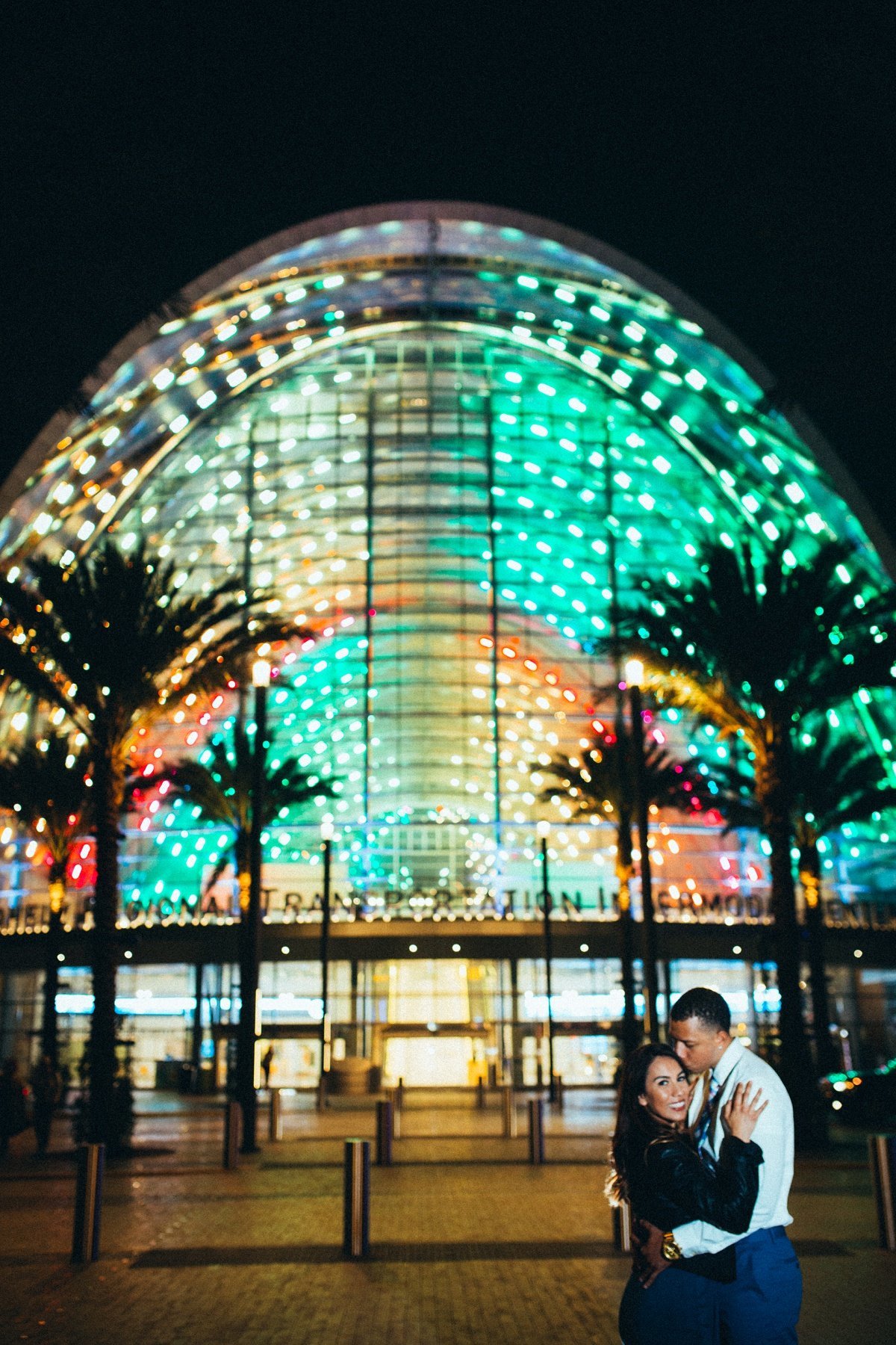 Groom to be kisses his Bride's head as she smiles for the camera at night in front of the colorful ARTIC Train Station at night