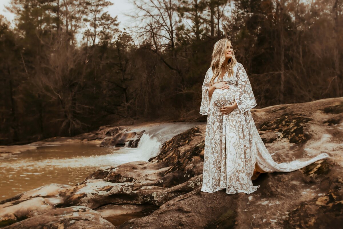 A pregnant woman in a lace dress stands on a rock near a small waterfall and forest, holding her belly and looking into the distance.