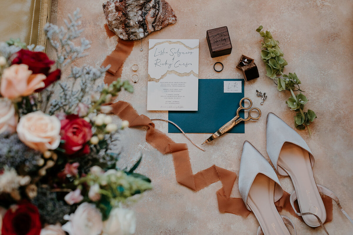 A flat lay of bridal accessories, including pointed-toe shoes, wedding rings, scissors, floral arrangements, and an invitation suite with "Leslie Salgueso and Ricky Carson," against a soft neutral background with ribbon accents.