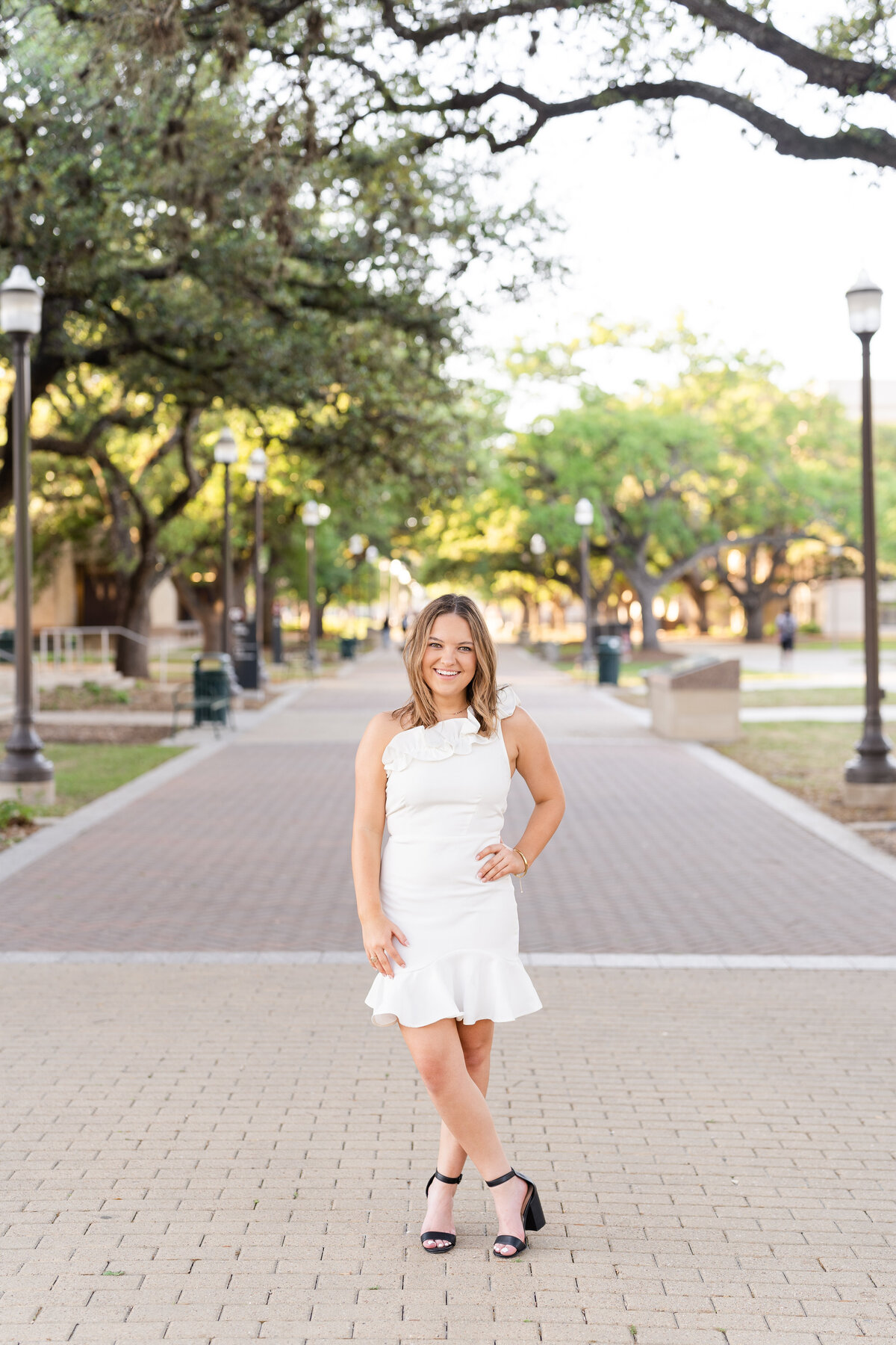Texas A&M senior girl standing with hand on hip smiling and wearing a white dress under the trees of Military Plaza