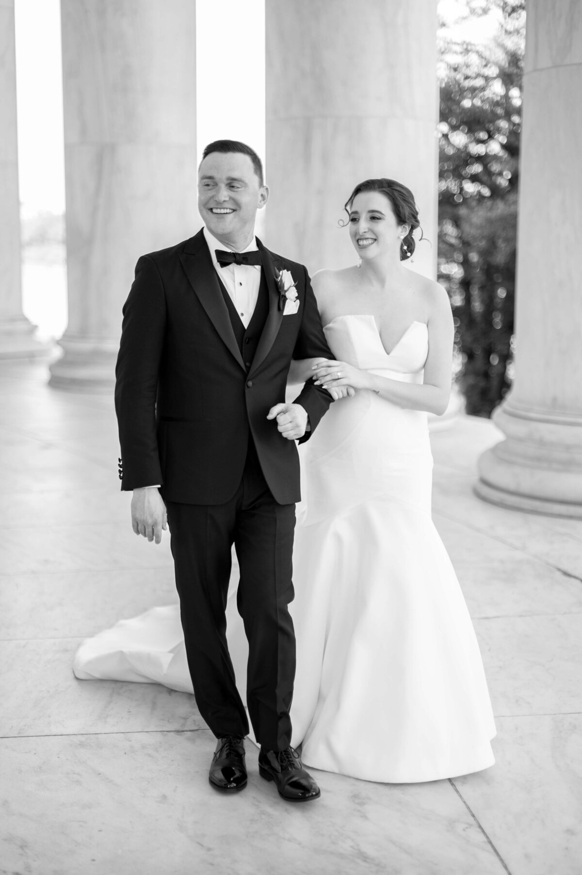 A bride in a strapless gown smiles as she links arms with a groom in a tuxedo. They stand between marble columns, exuding joy and elegance in a black and white setting.