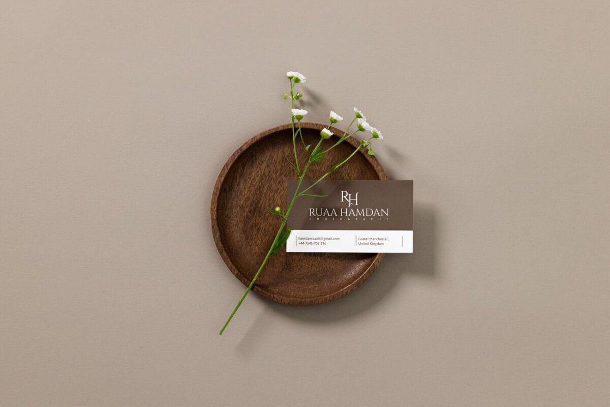 Brown wooden dish holding a single white baby’s breath flower and a professional photographer’s business card with clean minimalist design on a taupe background.