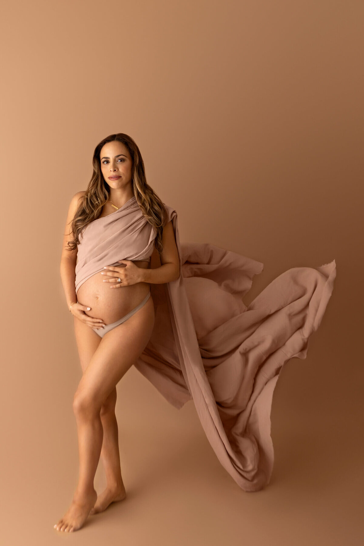 A pregnant woman stands gracefully against a warm beige background, draped in a flowing nude-colored fabric that wraps around her body and billows behind her. She wears matching nude underwear and cradles her baby bump with both hands, gazing softly at the camera. Her long, wavy hair cascades over her shoulders, adding to the elegance and serenity of the portrait.