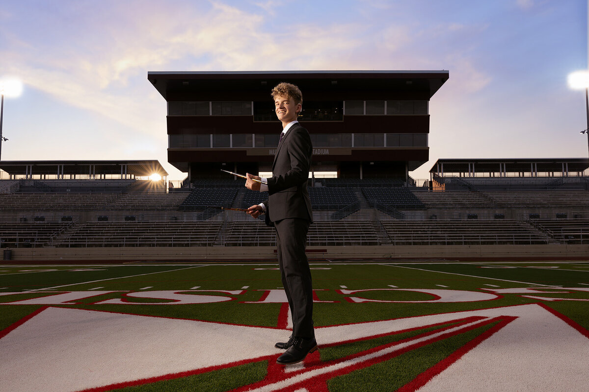 A high school senior smiles confidently while standing on the football field at Mustang Panther Stadium, holding drumsticks. Dressed in a sharp suit, he is framed by the empty stadium stands in this Dallas senior photography session at sunset.
