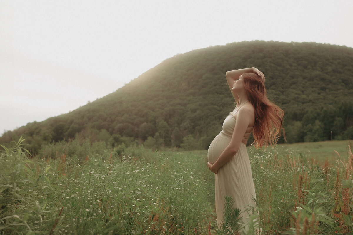 a maternity photo of woman with long red hair standing in an open field of wildflowers located in Finger Lakes NY