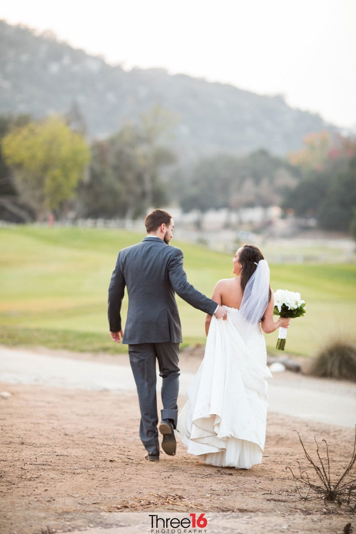 Groom carries his Bride's dress train as they walk towards the golf course