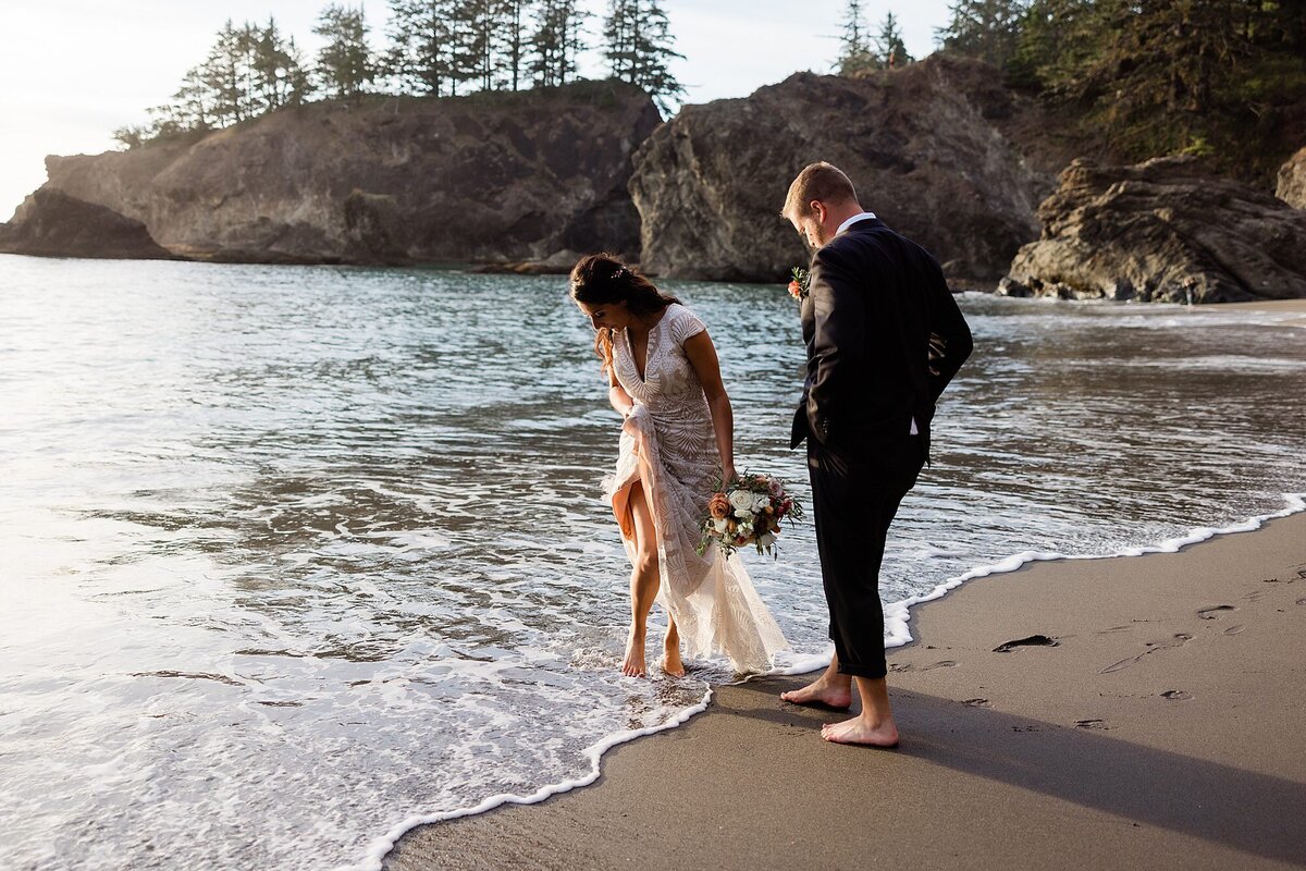 A bride and groom dip their toes in the Pacific ocean.