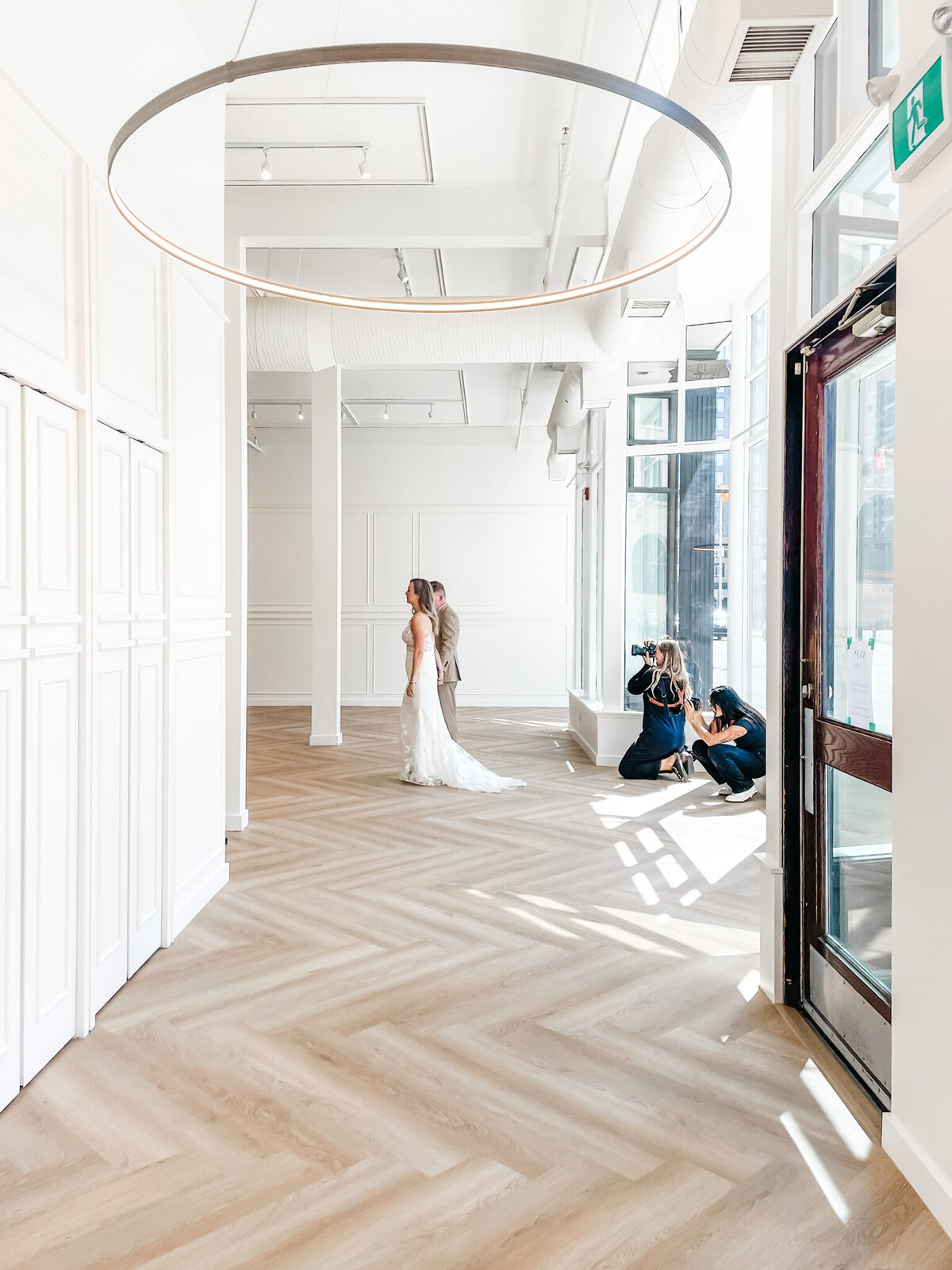 A spacious, modern event venue at Studios at Vic Park in downtown Calgary, featuring large windows with sheer white curtains, herringbone wood flooring, and contemporary furnishings. Bright natural light enhances the elegant and versatile space.