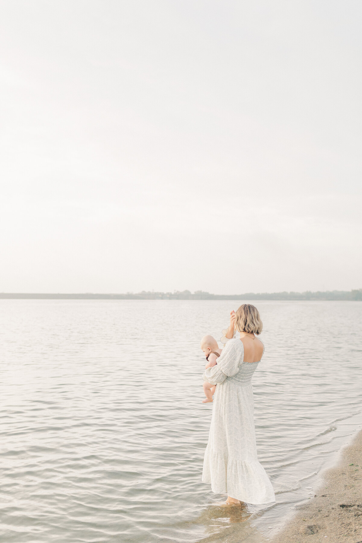 Mom stands at the edge of the lake holding her baby, captured during a calm 6-month milestone session in Kansas City.