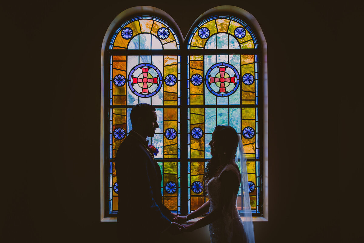 Indian christian bride and groom hold hands in front of stained glass windows inside arch angel michael orthodox church during the wedding in port washington ny