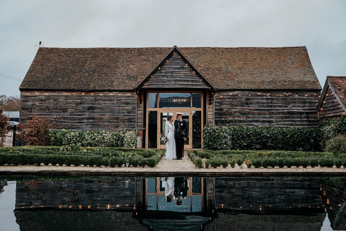 Bride and groom stand together infront of the barn at Silchester Farm with the pond infront of them