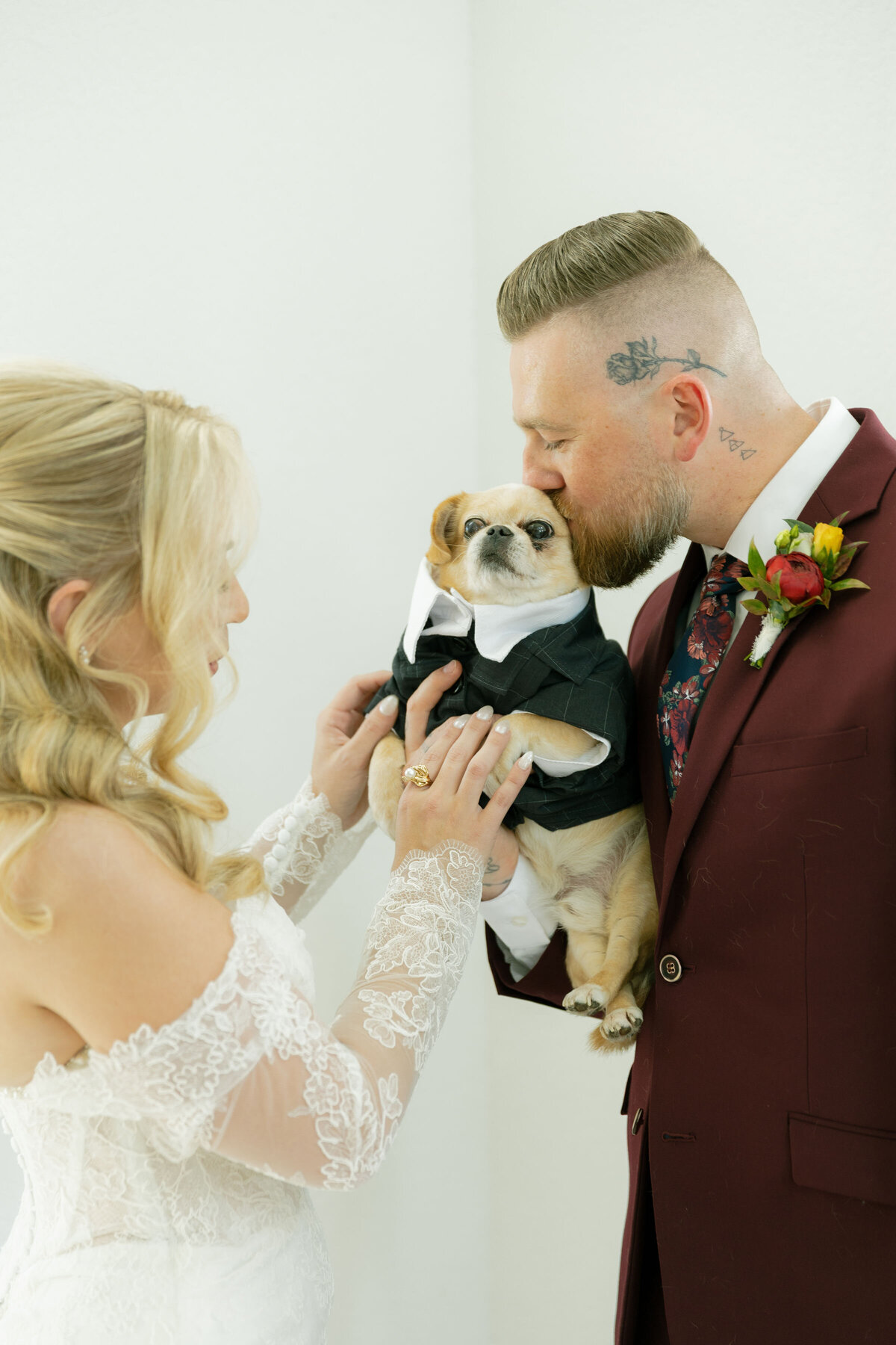 A bride and groom lovingly hold their small dog dressed in an adorable black wedding tuxedo during their getting-ready photos. The bride’s lace off-the-shoulder gown and the groom’s burgundy suit frame the dog as the groom gently kisses its head. A sweet and candid wedding moment capturing personality, warmth, and the couple’s cherished pet.