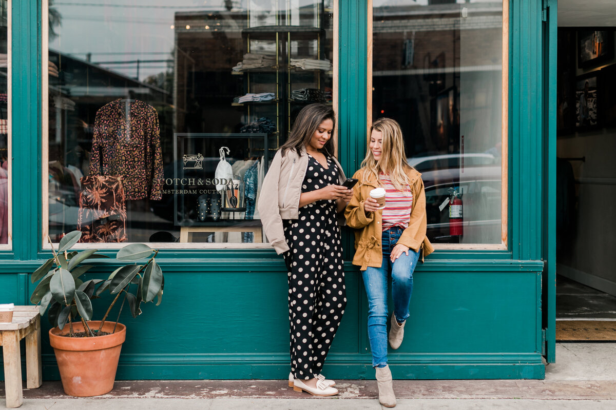 Co-Founders Andrea & Liz casually standing on a sidewalk smiling with their phone and coffee