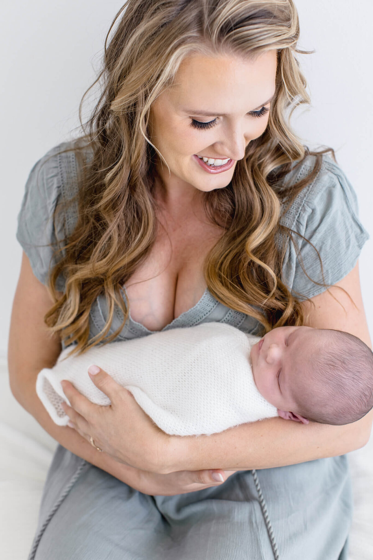 A woman with long, wavy hair smiles warmly as she cradles a sleeping newborn wrapped in a white blanket. She is wearing a light gray dress, and they are seated in a softly lit setting.