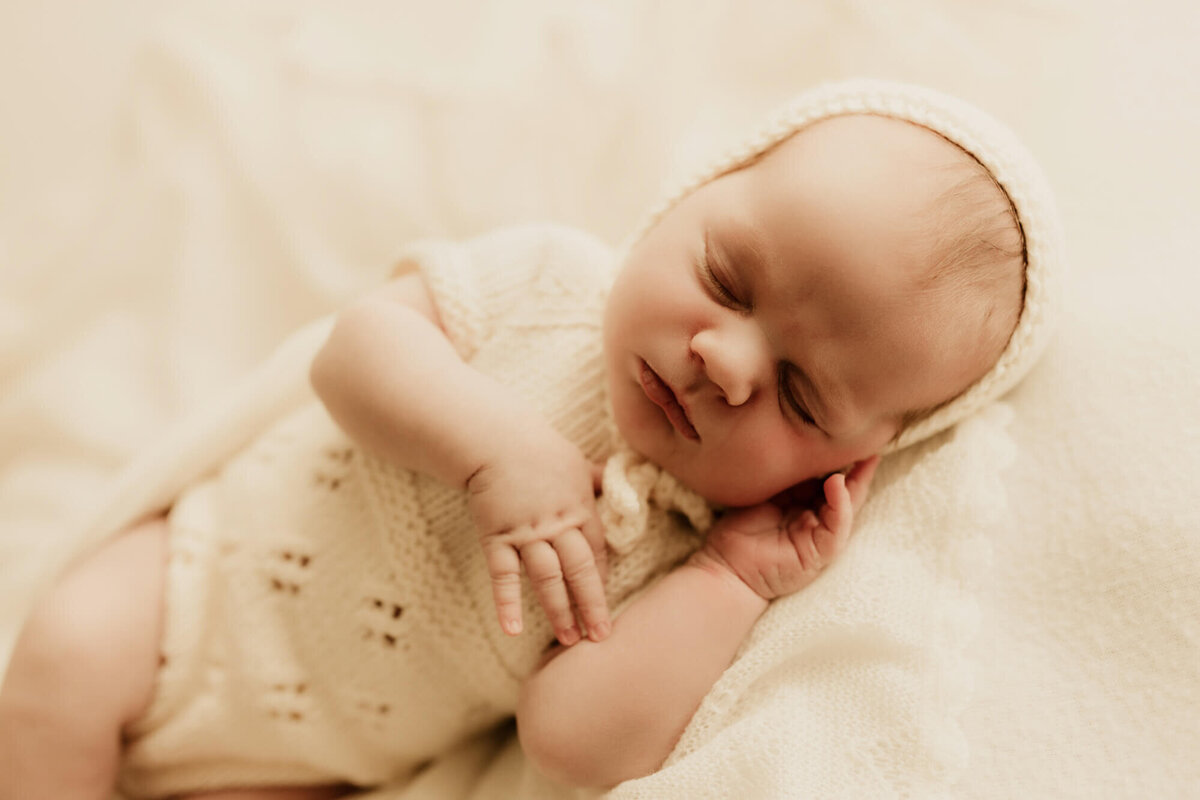 Newborn baby girl sleeping on her side while wearing a cream knit romper and matching bonnet, Edmond, Oklahoma.