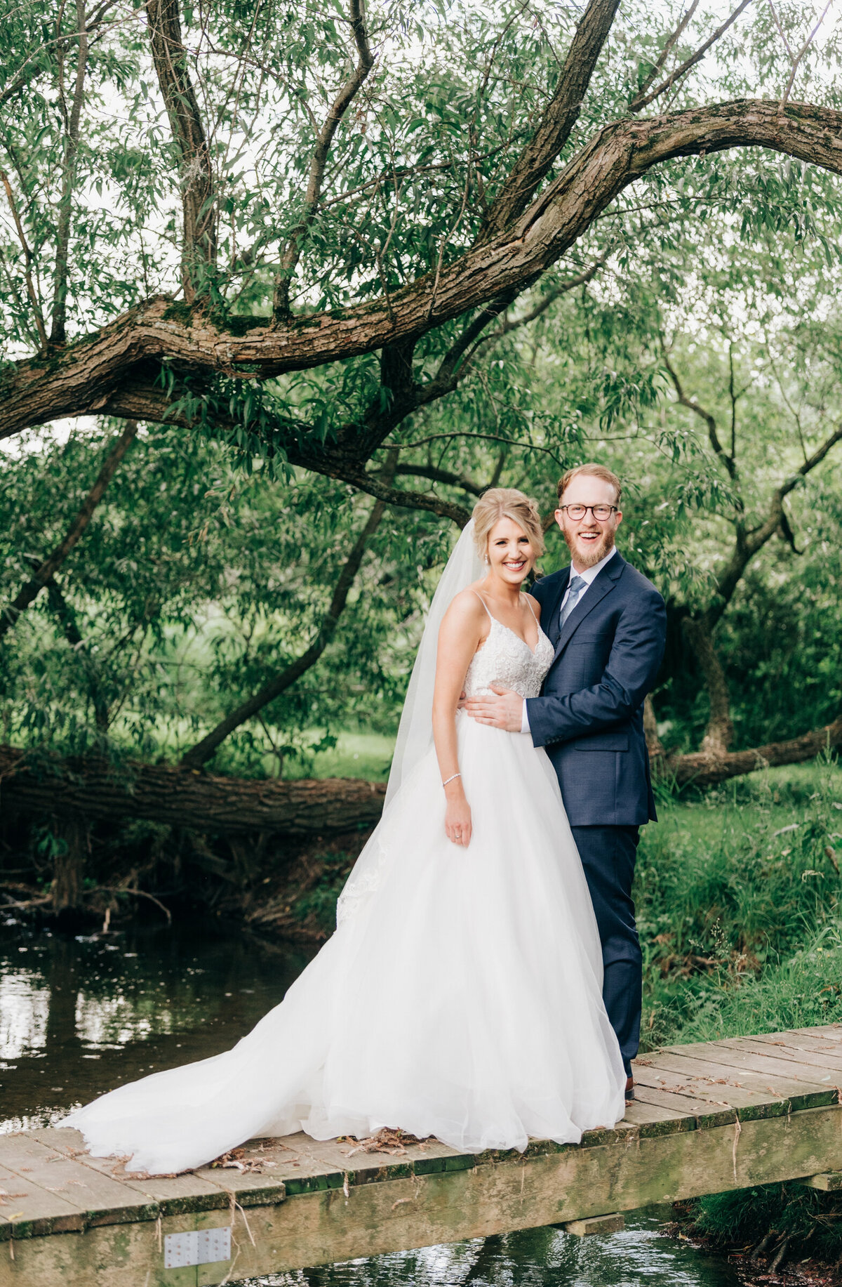 Elegant bride and groom stand on bridge under gorgeous willow tree at Willow Creek Barn photographed by Nova Markina
