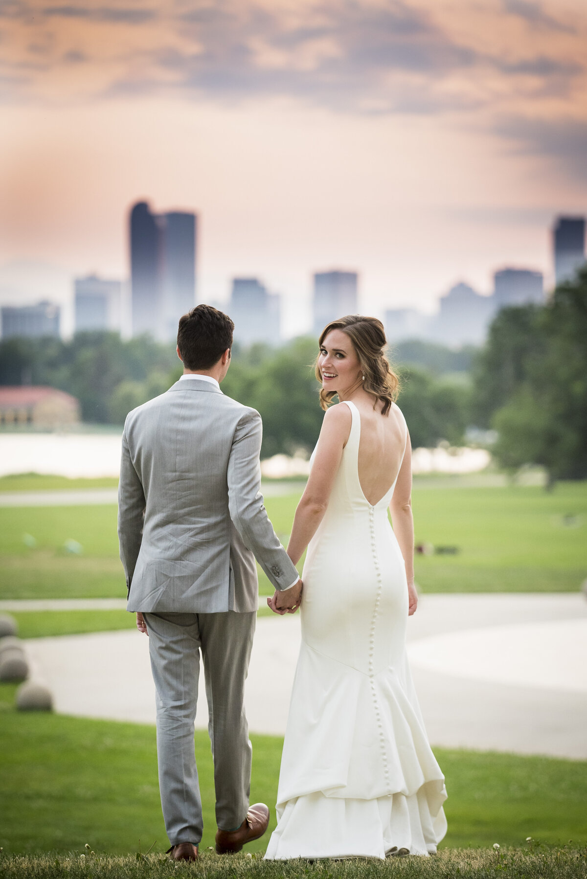 A bride and groom face away from the camera as the bride looks over her shoulder to smile with the Denver skyline in the background.
