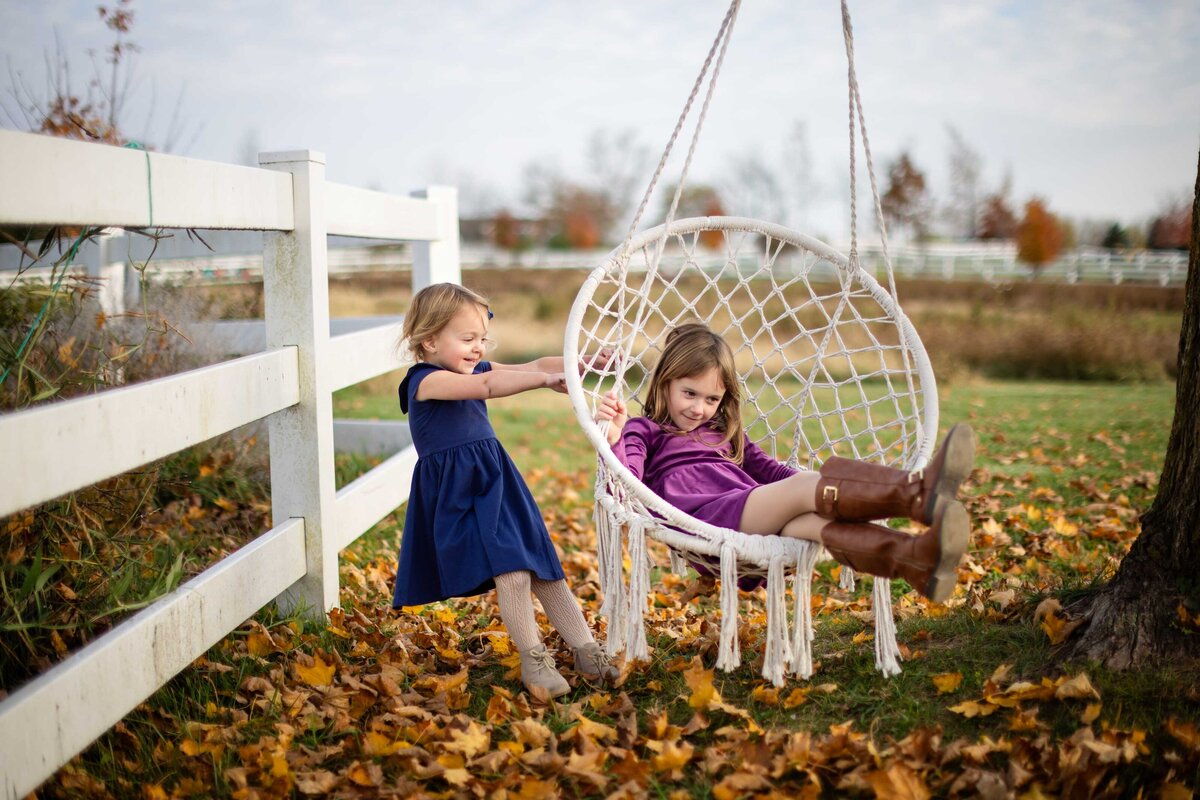 Two young girls enjoying a playful moment outdoors in Metro Detroit, one pushing her sister on a hanging white swing surrounded by fallen autumn leaves.