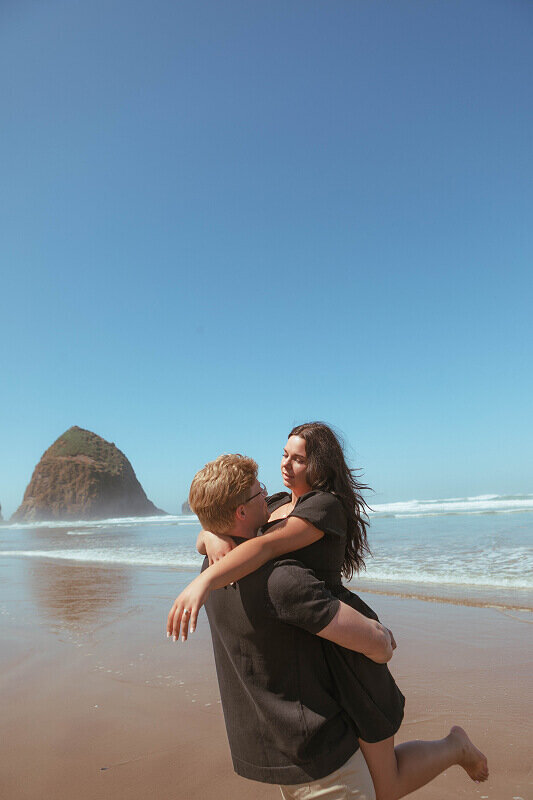 A man holds and twirls his fiancé on the beach in Oregon during their engagement shoot. 