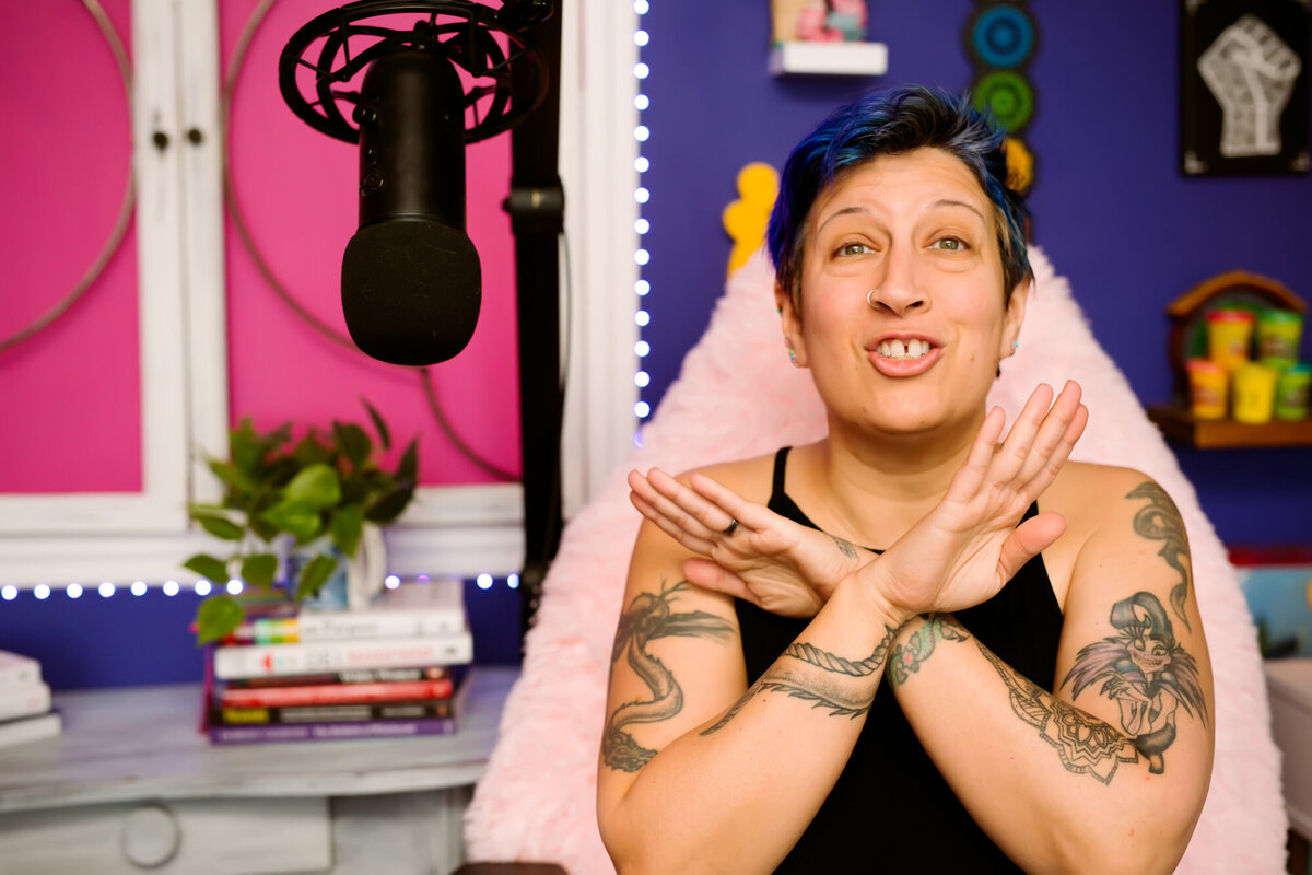 They/them podcaster sitting in front of a microphone doing a podcast with her hands crossed in front of her to show "stop shoulding yourself." posing for a South Shore MA branding photographer