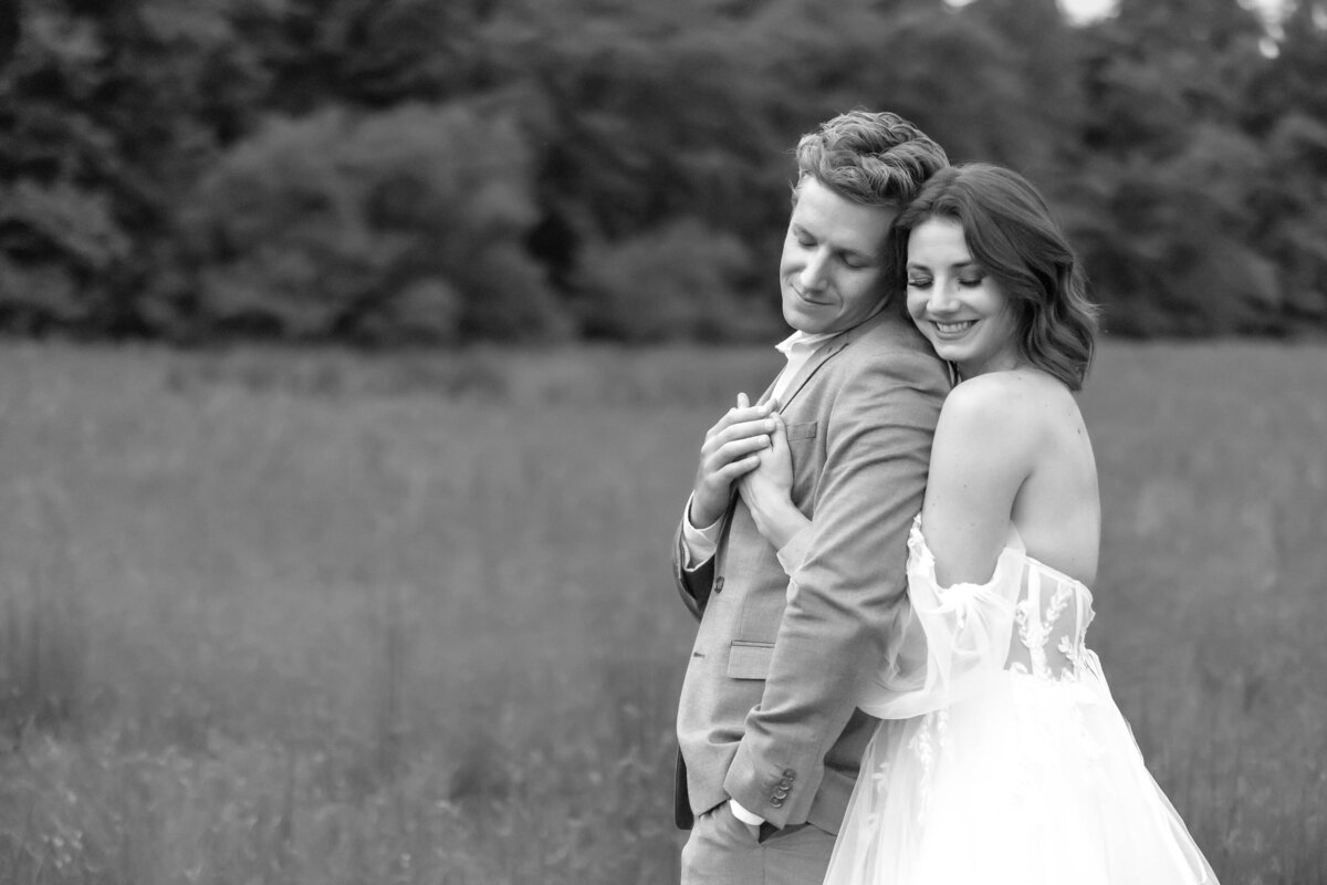 Experience the elegance of a black and white photo capturing a bride embracing her groom and resting her head against his, set in a serene field, symbolizing timeless love and connection.