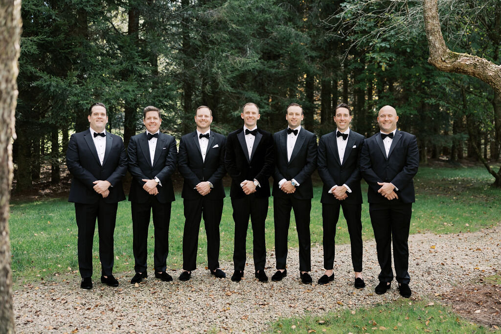 Groomsmen in classic black tuxedos pose outdoors under tall evergreens during a Highlands, North Carolina wedding.