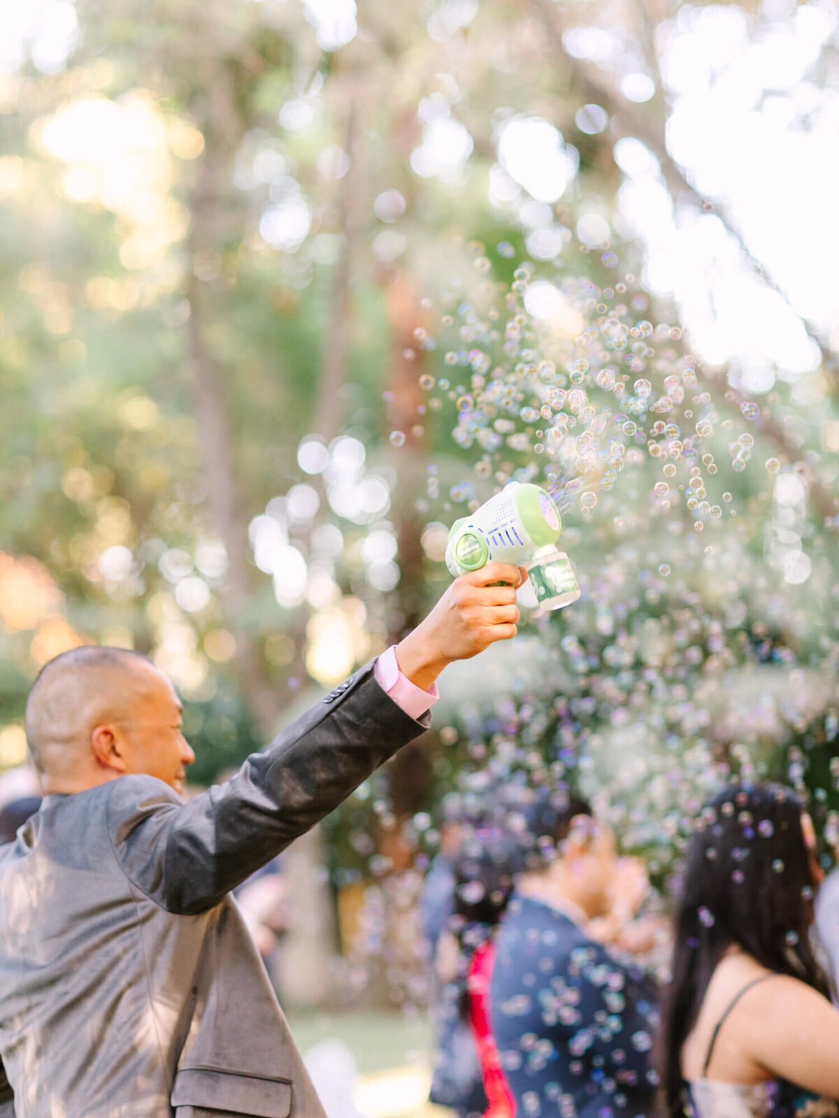 A guest in a suit joyfully uses a bubble machine outdoors. Bubbles fill the air against a backdrop of trees and crowd.