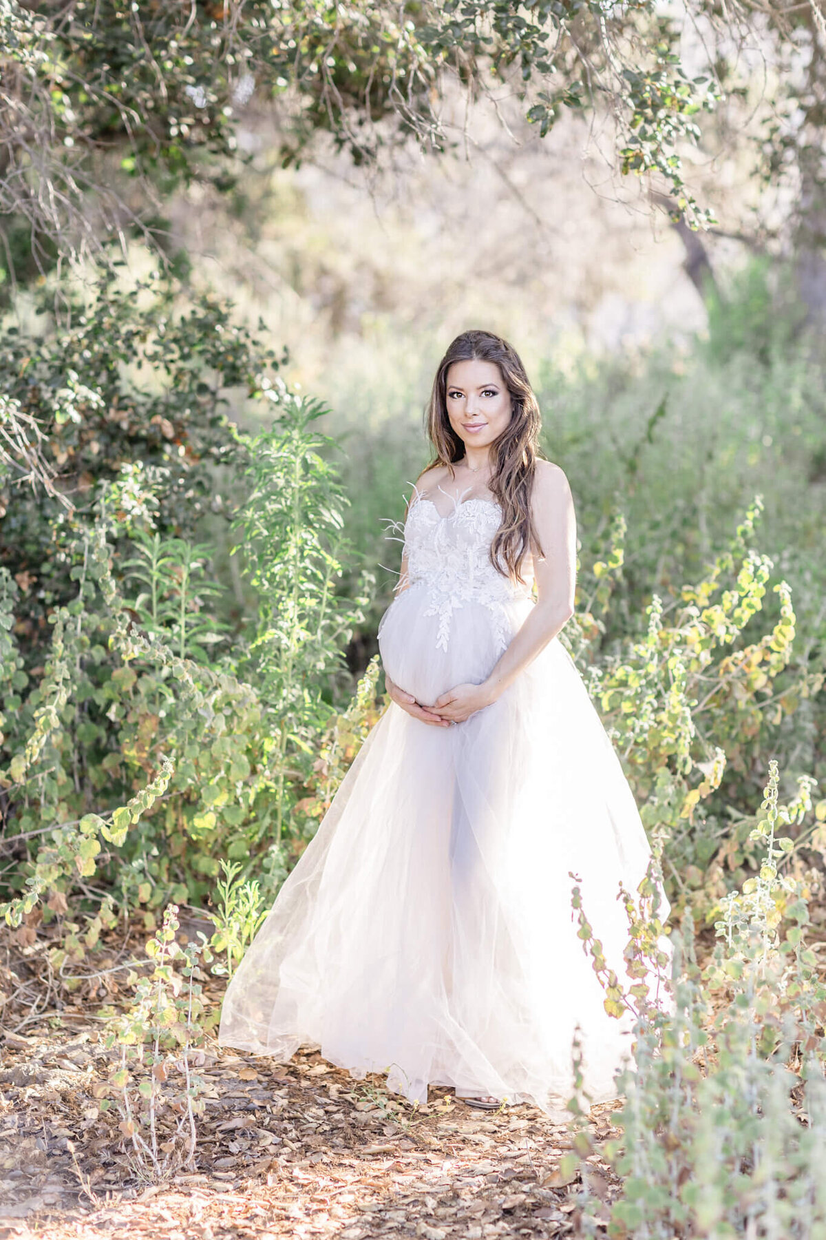 A pregnant woman in a white, flowing dress stands in a sunlit forest. She cradles her belly with both hands, surrounded by green foliage and dappled light filtering through the trees.