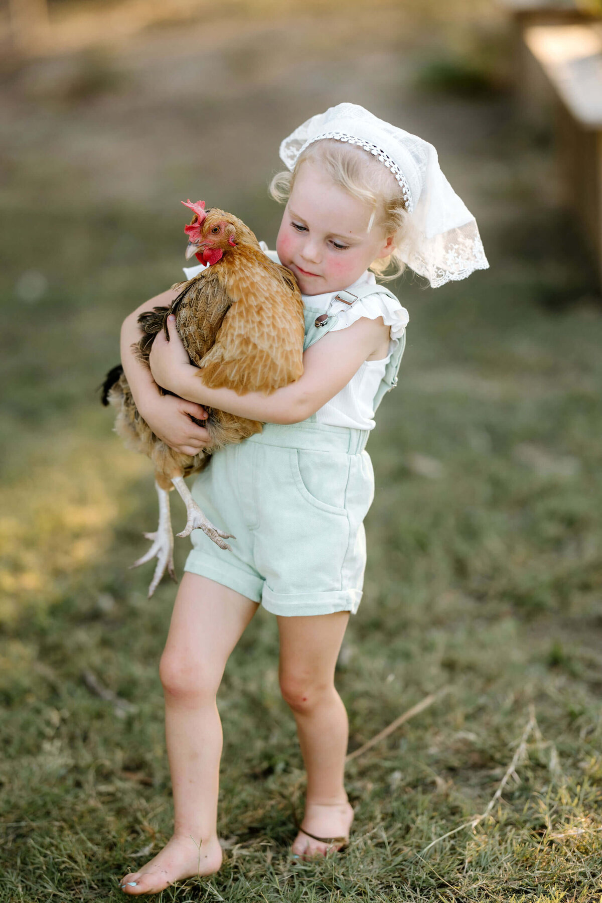 candid photo of 3 year old girl in green overalls and white bonnet carrying chicken on Hallsville, TX family farm
