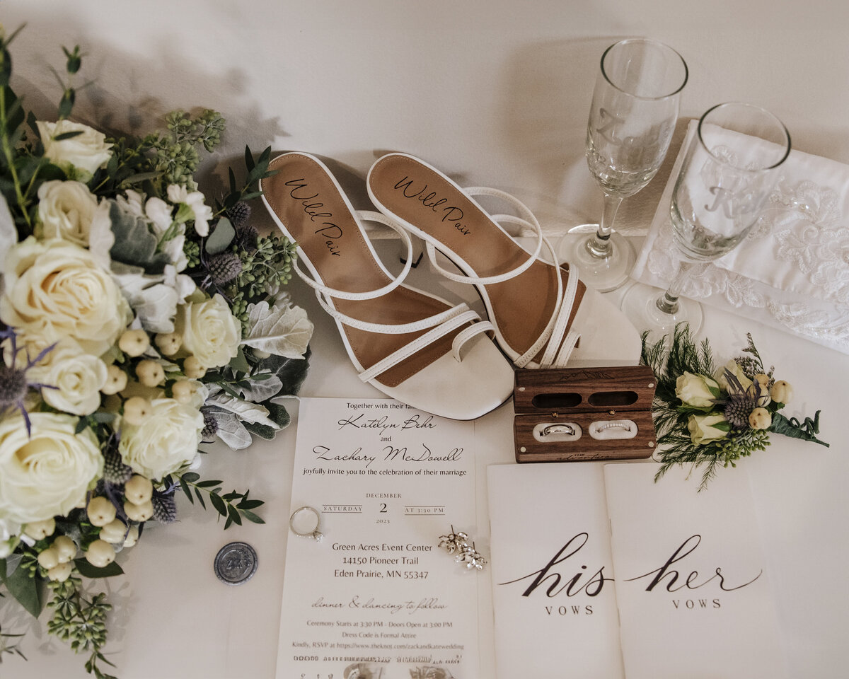 Elegant wedding accessories and keepsakes gracefully displayed, including bridal shoes, handwritten vows, invitation suite, rings in a wooden box, and delicate floral arrangements taken by jen Jarmuzek photography a Minneapolis wedding photographer